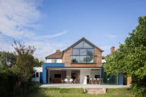 A modern two-story house with large glass windows, set in a garden with a lawn. The house features a blue and white exterior with an open patio area and visible interior furnishings.