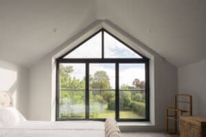 Bedroom with a triangular window showcasing a garden view. Room includes a bed, wicker basket, and wooden shelf.