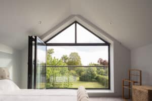 A bedroom with a large triangular window, open to a view of a garden. The room has a bed and wicker basket.