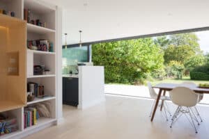 Modern open-plan kitchen and dining area with white walls, light wood floors, a bookshelf, and large glass doors opening to a garden.