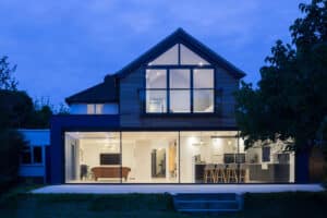 Modern two-story house with large glass windows, lit from inside. Open living space visible through windows, surrounded by trees, under a dusky sky.