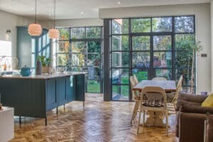 A modern kitchen with teal cabinets, a wooden dining table, and large windows overlooking a garden.
