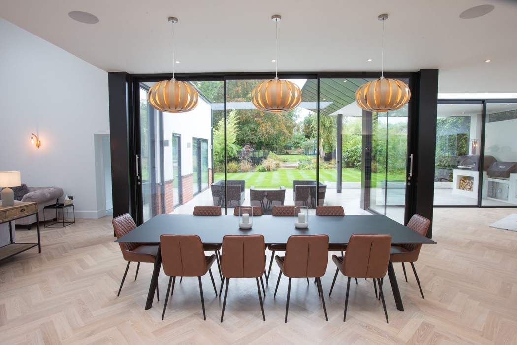 Modern dining room with a black table and eight brown chairs. Three pendant lights hang above. Glass doors open to an outdoor patio and garden. Light wood flooring throughout.