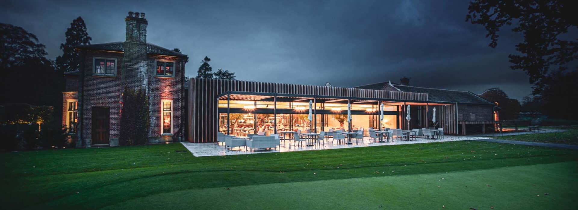 Modern building with large glass windows and sleek sliding aluminium doors, complemented by inviting outdoor seating, set against an evening sky on a grassy area.