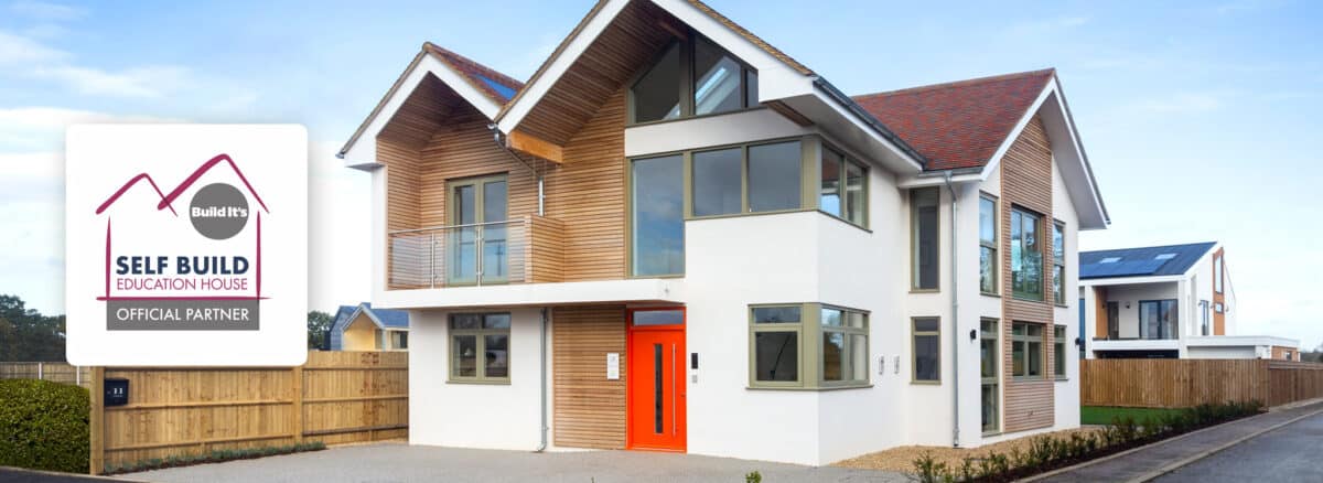 Modern two-story house with wooden paneling, large windows, and an orange front door complemented by sleek slimline sliding doors. A sign reads "Build It, Self Build Education House, Official Partner.