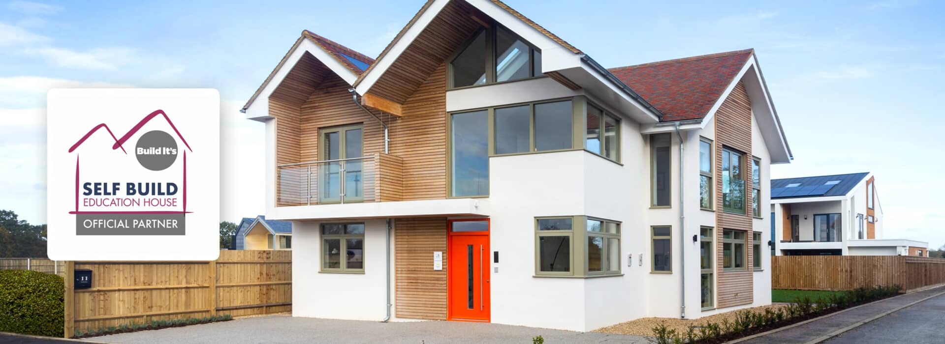 Modern two-story house with wooden paneling, large windows, and an orange front door complemented by sleek slimline sliding doors. A sign reads "Build It, Self Build Education House, Official Partner.