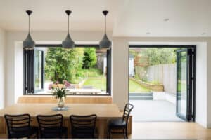 The dining area features a wooden table and black chairs under pendant lights. Large aluminum sliding windows and open doors provide a seamless view of the lush garden with a patio and outdoor seating.