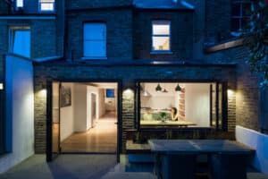Brick house with large bifold windows, patio, and outdoor dining area at dusk. The interior is visible through the expansive aluminium frames, showing people sitting and dining.