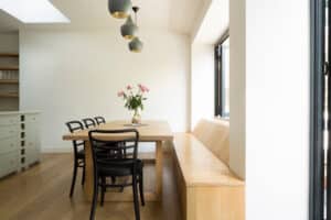 A modern dining area with a wooden table, black chairs, and a built-in bench sits gracefully beside sleek aluminium sliding windows. Three pendant lights hang above, and a vase with pink flowers adorns the table, perfectly blending elegance and contemporary design.