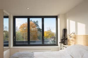 The bedroom features a large aluminum-framed sliding window showcasing views of trees and a construction crane. The bed is dressed in crisp white linens, and a telescope stands ready by the window.