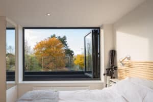 The bedroom features a bifold window that perfectly frames the view of autumn trees. Inside, you'll find a cozy bed with white linens and a sleek silver lamp on the headboard, enhancing the tranquil atmosphere.