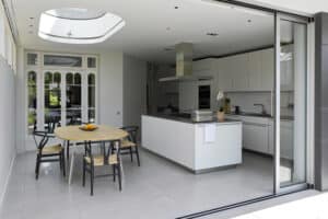 Modern kitchen with white cabinets, central island, and a skylight. A round dining table with chairs is nearby, and large glass pocket sliding doors open to a garden.