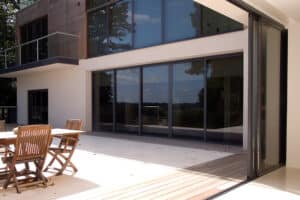Modern house exterior featuring expansive glass windows and sleek pocket sliding doors. Wooden patio furniture on the deck enhances the seamless indoor-outdoor flow.