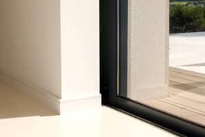 A corner of a room with white walls and a large glass pocket sliding door. The sleek door easily opens to reveal an outdoor wooden deck with greenery in the distance.