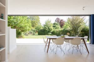 Modern dining area with a wooden table and white chairs, seamlessly connected to a lush green garden through large pocket sliding glass doors.