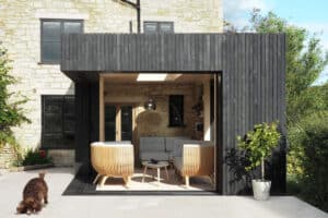 A modern outdoor seating area features wooden armchairs and a coffee table, all elegantly enclosed by dark wooden panels and pocket sliding doors. A cat happily walks nearby on a sunny day, adding life to the serene scene.