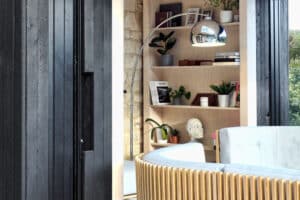 A cozy modern living room featuring pocket sliding doors, a curved light wood accent chair, a tall arc floor lamp, and shelves with books and plants against a stone wall.