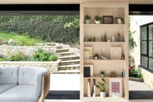 Modern living room with beige shelving unit displaying books, plants, and decor. Gray sofa on the left. Pocket sliding doors open to reveal a garden with stone steps and lush greenery.