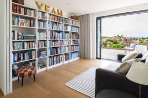 Modern living room with a large bookshelf, a gray couch, a round footstool, and an area rug. Pocket sliding doors open to a garden. The word "YEAH" is displayed on top of the bookshelf.