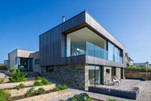 Modern two-story house with large glass windows, pocket sliding doors, stone and wood exterior, and a patio. Raised garden beds are in the foreground under a clear blue sky.