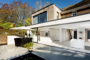Modern house with large bifold windows, an outdoor patio featuring a round dining table and umbrella, and an open kitchen area. Surrounded by trees under a clear sky.
