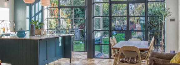 A bright kitchen and dining area features heritage doors that open to a garden. Inside, there's a wooden dining table, chairs, and a kitchen island with a teal teapot. Large windows flood the space with natural light, creating an inviting atmosphere.