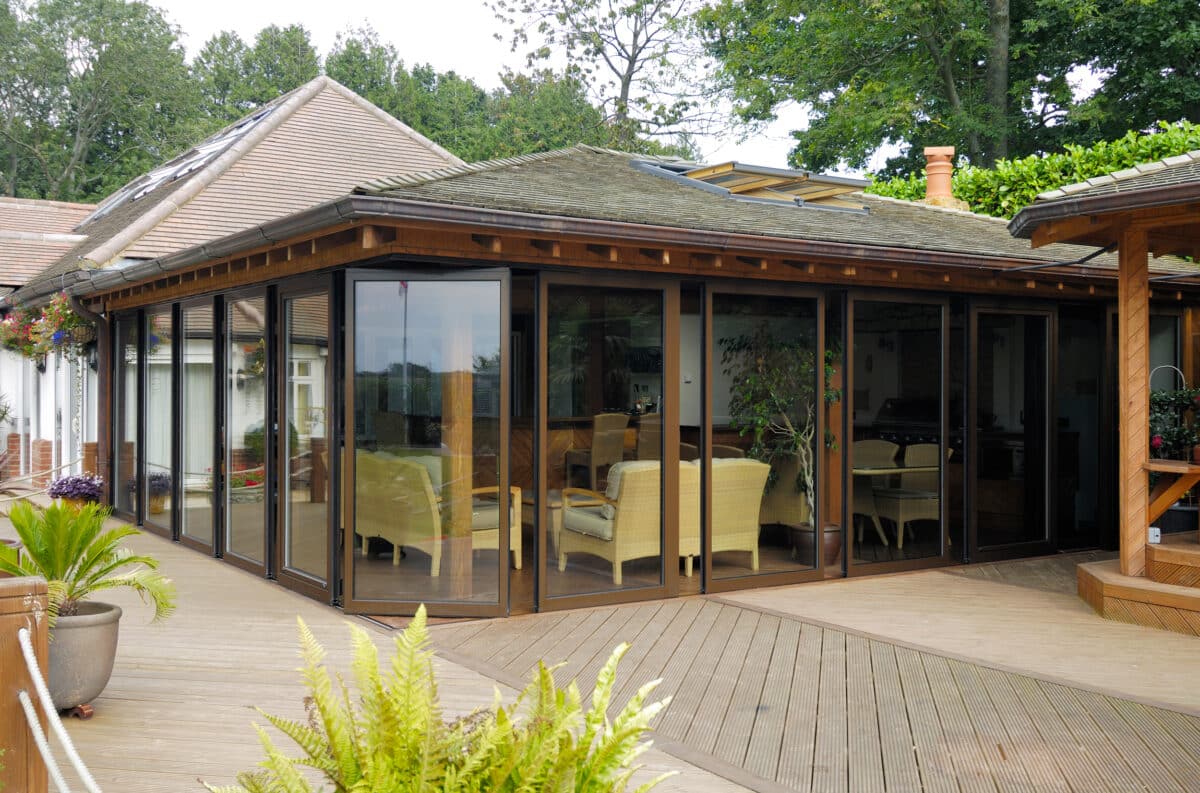 A modern sunroom with large glass windows, slide and stack doors, wicker furniture, and wooden decking surrounded by lush plants and trees.