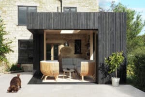 Modern outdoor living space with sliding aluminium doors leading to a wooden structure. Two chairs and a sofa create a cozy setting, while a potted plant and a small dog enjoy the stone patio. The background features a stone building surrounded by lush greenery.