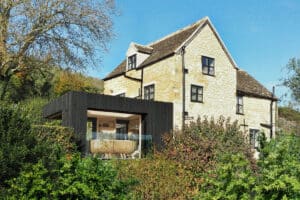 Stone house with modern black extension, featuring large windows and sliding aluminium doors, surrounded by trees and greenery under a clear blue sky.