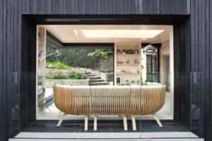 Contemporary interior featuring a large wooden bench facing sliding aluminium doors, which open to a garden with stone steps and lush greenery. Shelves with decor are visible in the background, enhancing the modern aesthetic.