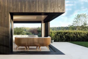 A modern outdoor patio with wooden slat furniture, open sliding aluminium doors, and a view of greenery and hills.