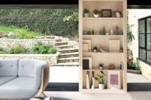 A cozy room with a wooden bookcase holding plants and decor items. A cushioned chair sits nearby, framed by sliding aluminium doors that reveal a garden visible through large windows in the background.