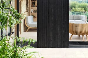 Modern living space with light wooden furniture separated by a dark vertical panel. Potted plants are visible in the foreground, while large sliding aluminium doors reveal a garden view.