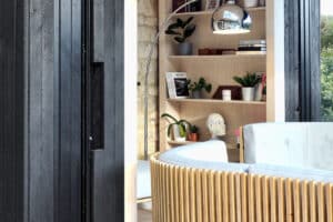 Modern living room with a curved wooden sofa, a floor lamp, and a bookshelf adorned with plants and decor. Sliding aluminium doors open to reveal black wall paneling partially visible on the left.