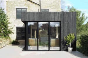 A modern addition with sliding aluminium doors complements the stone building; interior features chairs and a table, seamlessly merging with the greenery outside.