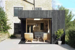 Modern outdoor living space with wicker chairs and a sofa under a black wooden awning, attached to a stone building. Sliding aluminium doors seamlessly connect the indoors to this serene oasis, enhanced by lush greenery surrounding the area.