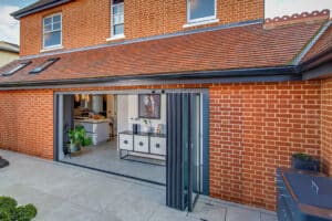 Modern brick house featuring sleek slide and turn doors that open to a kitchen area. The outdoor space boasts light-colored tiles and a few plants, enhancing its inviting ambiance.
