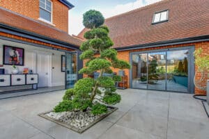 Courtyard with a sculpted tree centerpiece, surrounded by red-brick buildings with large windows and slide and turn doors, all beneath a charming tiled roof.