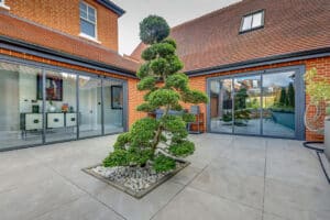 Modern courtyard with a neatly trimmed bonsai tree in a square planter, surrounded by red brick buildings and large slide and turn glass doors.