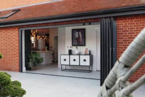 Open slide and turn doors reveal a modern living space with a console table and artwork. The kitchen is visible in the background, while the brick exterior and patio seating are featured in the foreground.