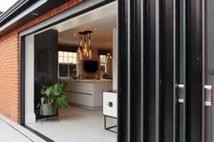 Open slide and turn doors revealing a modern kitchen with a potted plant, island counter, and hanging light fixture.