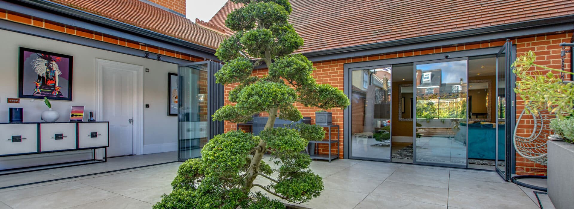 A modern courtyard features a manicured tree, surrounded by sleek slide and turn doors that lead to the kitchen and living area. A framed artwork and decorative cabinet adorn the left wall, enhancing the serene ambiance.