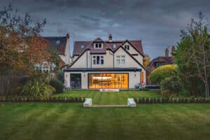 A two-story house with a large, modern extension featuring sleek slide and turn doors, a well-maintained lawn, and exquisite landscaping, all under a cloudy sky.