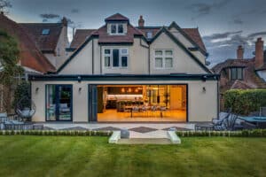 A suburban house features large slide and turn glass doors that open to a patio and garden. The ground floor interior is warmly lit, revealing a dining area and kitchen.