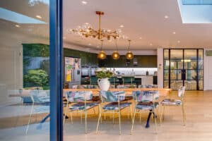 Modern kitchen and dining area featuring a large wooden table, patterned chairs, gold light fixtures, and green cabinets. Slide and turn doors elegantly open to reveal the outdoor space.