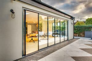 Modern house exterior with impressive slide and turn doors revealing a stylish indoor living area. The patio features a smooth tiled surface and elegant outdoor lighting.