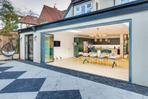 Spacious home with slide and turn doors opening to a modern kitchen and dining area, featuring a large table and accent lighting, leading onto a checkered stone patio.