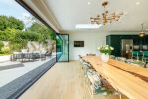 Spacious open-plan dining area featuring a large wooden table, floral chairs, and modern lighting. Slide and turn doors seamlessly open to a patio with outdoor seating and a garden view.
