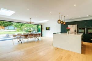 Spacious kitchen with a large island, green cabinets, pendant lights, and a dining area. Floor-to-ceiling slide and turn doors open to a garden, illuminating the space with natural light.