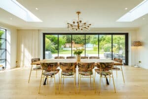 A spacious dining room with a long table and 10 chairs. Slide and turn doors open to a garden, enhancing the airy feel. The room features a chandelier and a floor lamp, with skylights on the ceiling.
